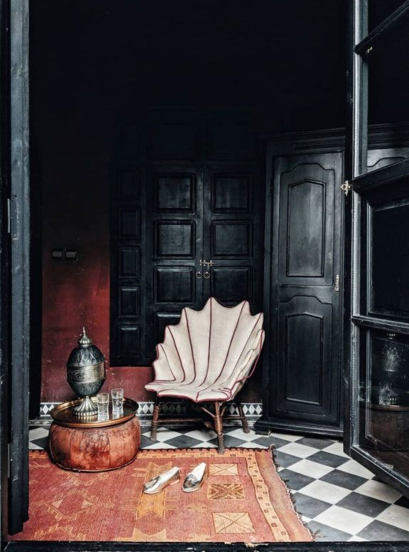 Dark interior with black carved woodwork, deep red walls, geometric tiled floor, vintage chair and warm-toned woven rug.