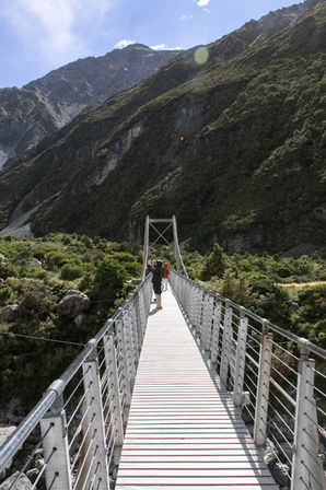 Besucher überqueren die Hängebrücke am Hooker Valley Track mit steilen Berghängen im Mount Cook National Park.