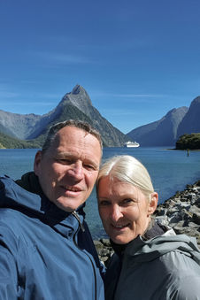 Paar beim Selfie vor dem Milford Sound mit markanter Bergkulisse und Kreuzfahrtschiff im Hintergrund