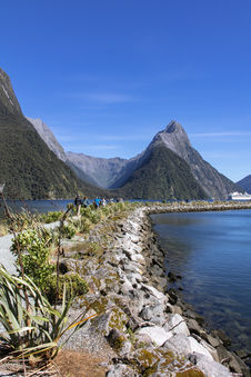 Spazierweg am Ufer des Milford Sound mit Blick auf markante Berge und Kreuzfahrtschiff in der Ferne