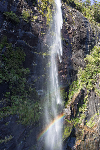 Wasserfall am Milford Sound mit leuchtendem Regenbogen in der Gischt vor einer steilen Felswand.