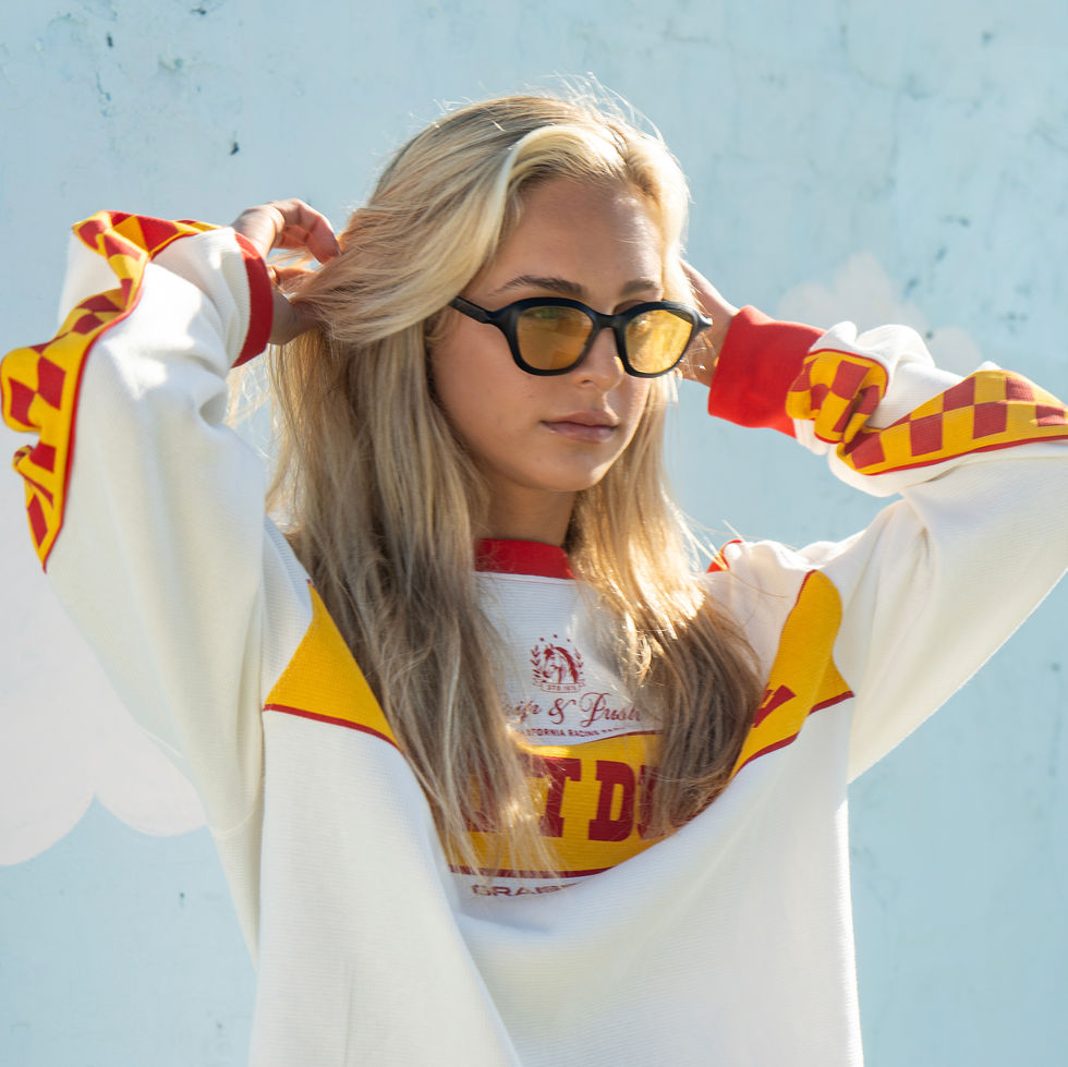 Blonde woman wearing black Orel sunglasses with yellow lenses and a sporty sweater outdoors.