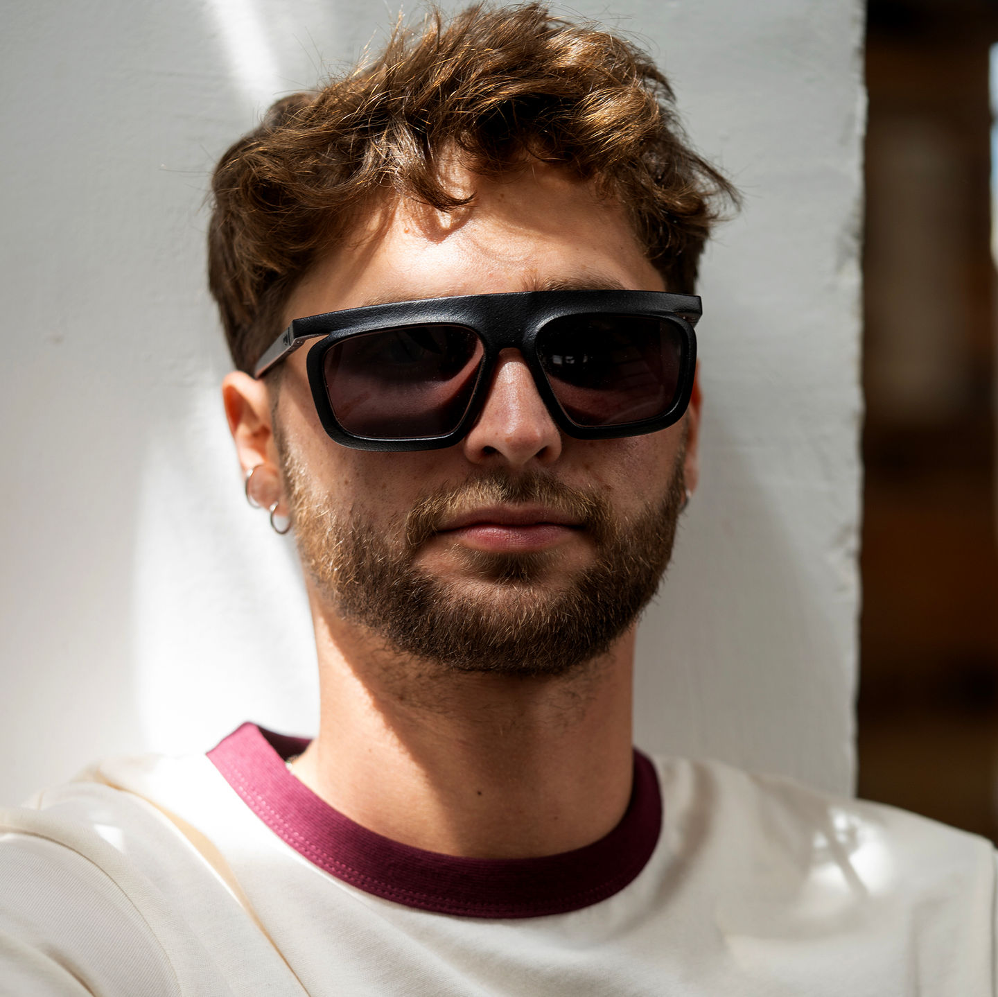 Bearded man wearing black Flat Top sunglasses with dark tinted lenses and a white T‑shirt with a burgundy collar