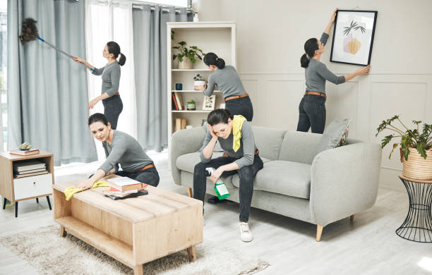 A woman in a gray shirt does multiple house chores in a living room; dusting, cleaning windows, arranging books, hanging a frame, and more.