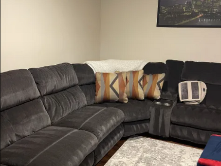 Dark gray sectional with patterned pillows and a white throw in a beige living room, with a framed city skyline above, cozy and calm