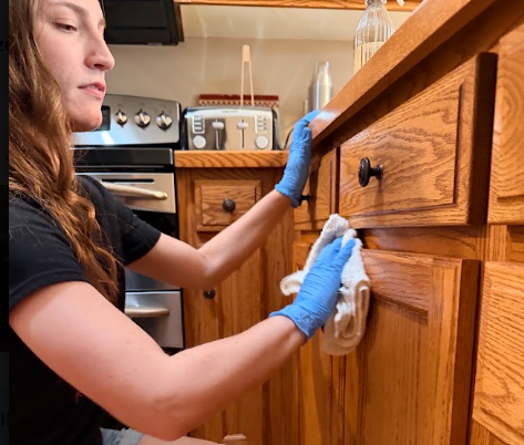 Sparkling Homes cleaner in blue gloves wipes a wooden kitchen cabinet with a cloth, focused in a warm home kitchen.