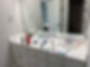 Messy white bathroom vanity with toiletries, toothbrushes, cups, and spray bottles scattered under a large mirror; toilet visible in doorway.