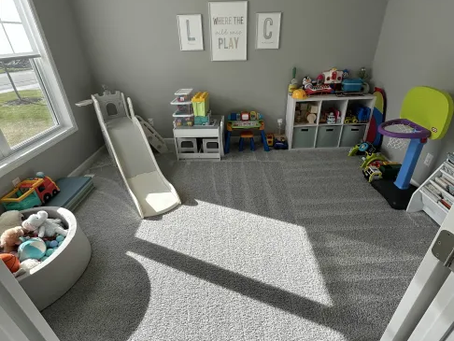 Bright playroom fresh vacuumed  with gray walls and carpet. Toys are neatly organized with a small slide and ball pit. Sunlight streams in, adding warmth.