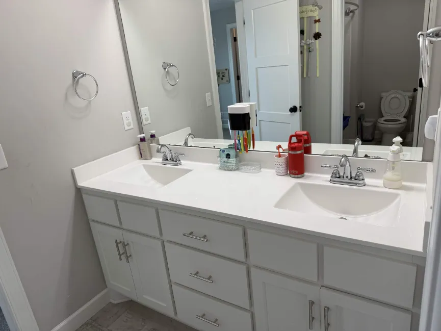 Bright bathroom vanity with double sinks, mirror, and toiletries; open door reveals a toilet room in the background.