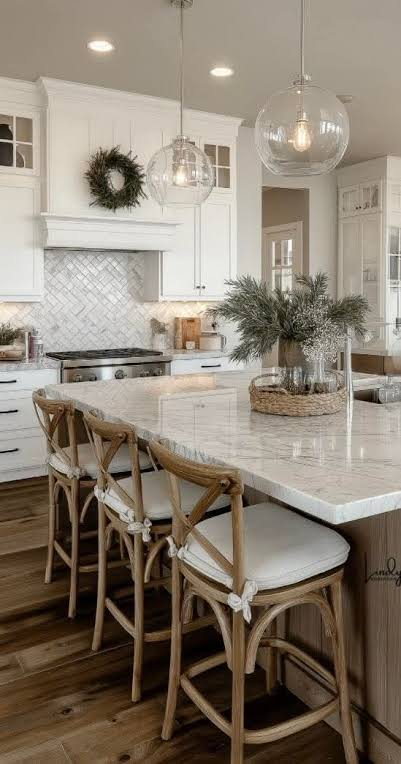 Bright modern kitchen with white cabinets, marble island, woven chairs, pendant lights, and a wreath above the stove.