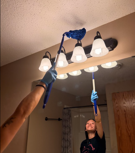 Two gloved sparkling homes cleaner cleaning a bathroom light fixture with long-handled mops, one smiling in the mirror under warm lights.