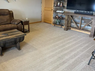 Living room with brown recliner, wooden table, large TV on rustic wood wall. Fish decor, beige carpet, and door in background. Cozy vibe.