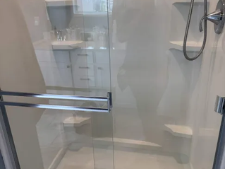 Glass shower with chrome fixtures and built-in shelves in a white bathroom, reflecting a person taking the photo.