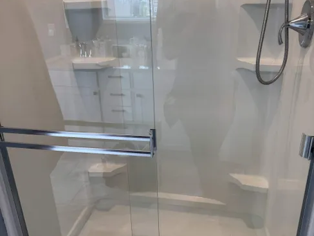 Glass shower with chrome fixtures and built-in shelves in a white bathroom, reflecting a person taking the photo.