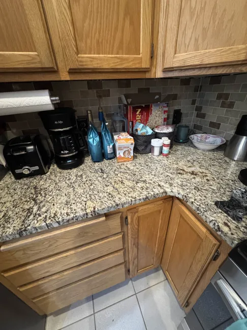 Granite kitchen counter with toaster, coffee maker, blue bottles, cereal box, spices, and bowls. Wood cabinets and tile backsplash.