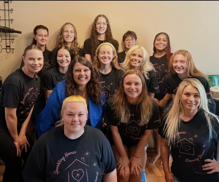 Group of smiling women in black Sparkling Homes Cleaning Services shirts pose together indoors against a beige wall.