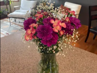 Bright bouquet of purple and pink flowers in a glass vase on a kitchen counter, with a gray living room and chairs behind.