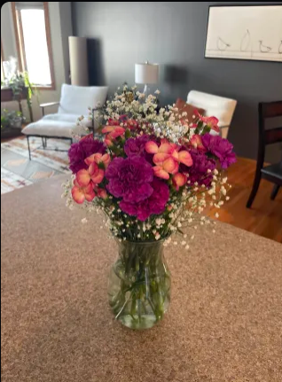 Bright bouquet of purple and pink flowers in a glass vase on a kitchen counter, with a gray living room and chairs behind.