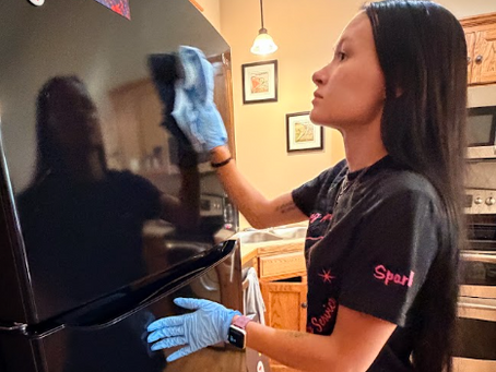 Sparkling homes cleaner in blue gloves wipes a black refrigerator in a warm kitchen, focused and reflected in the glossy door.