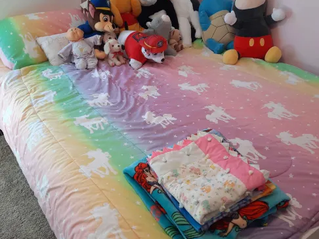 Colorful bed with unicorn-patterned blanket, plush toys including a panda and mouse lined up against the wall, folded clothes at the foot.