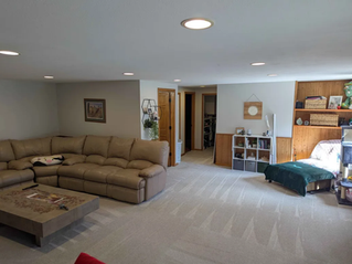Spacious living room with beige sectional sofa, wooden coffee table, shelves, and artworks. Sunlit, cozy atmosphere with neutral tones.
