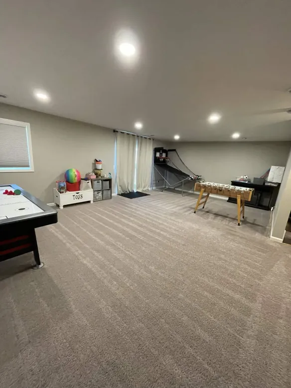 A spacious game room with a foosball table, air hockey, toys, and basketball setup. Beige carpet, neutral walls, and bright lighting.
