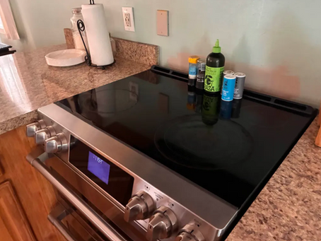 Stainless electric stove with black cooktop on a granite counter, seasoning bottles on top, and a lit blue clock display in a tidy kitchen.