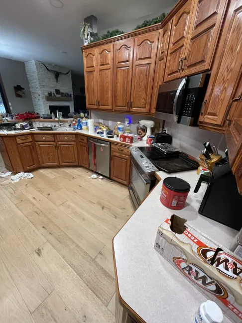Messy kitchen with wood cabinets and light countertops. Items like A&W box, utensils, and cans scattered. Fireplace visible in the background.
