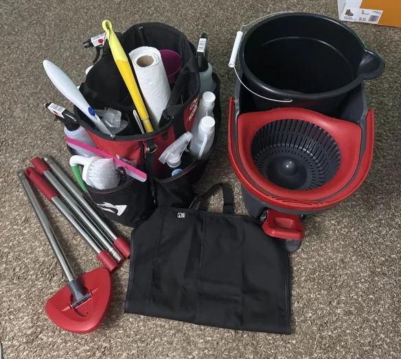 Cleaning supplies on carpet, featuring buckets, handles, brushes, and spray bottles in red and black. Neatly arranged, ready for use.