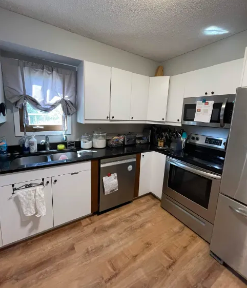 Bright kitchen with white cabinets, black counters, stainless steel appliances, a window over the sink, and papers on the microwave/dishwasher.