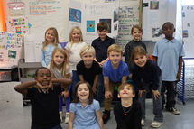 A class of elementary aged boys and girls, standing in a group posing, within a classroom.
