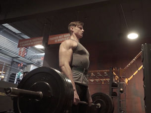 Muscular man in gym holding a heavy barbell, with Dutch safety signs hanging above in dim industrial lighting.