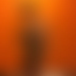 Muscular man takes a mirror selfie in a black cap and gray tank top against a bright orange wall, wearing earbuds.