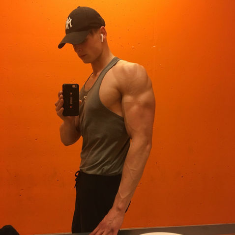 Muscular man takes a mirror selfie in a black cap and gray tank top against a bright orange wall, wearing earbuds.
