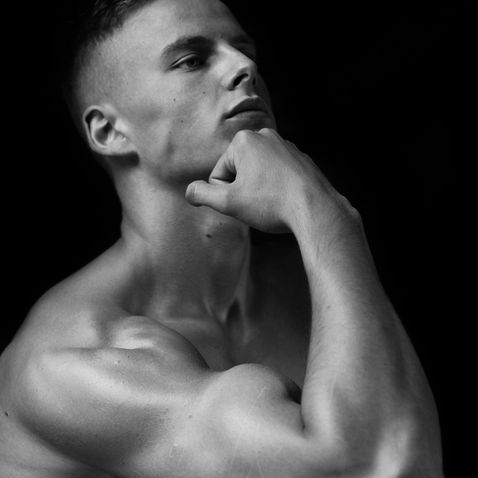 Black-and-white portrait of a shirtless young man with hand on chin, gazing upward against a dark background.