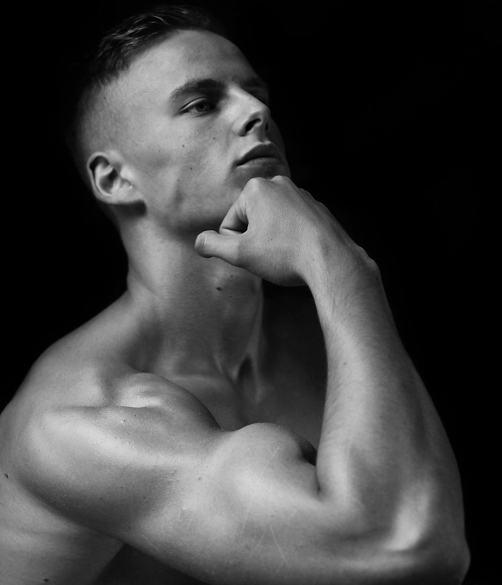 Black-and-white portrait of a shirtless young man with hand on chin, gazing upward against a dark background.