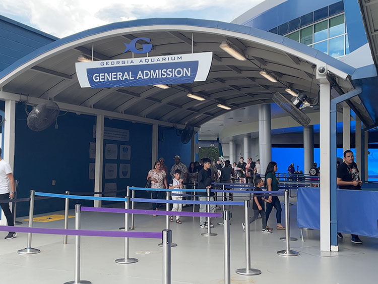 People entering Georgia Aquarium through General Admission. Blue and gray structure, signs overhead. Mood is busy and lively.