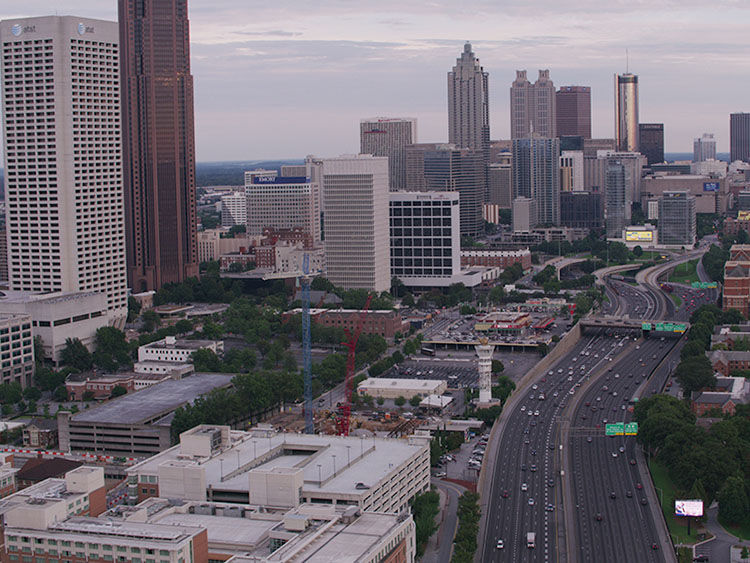 Atlanta City skyline with tall buildings, highway full of cars under cloudy sky, construction cranes visible, and greenery dotting the landscape.