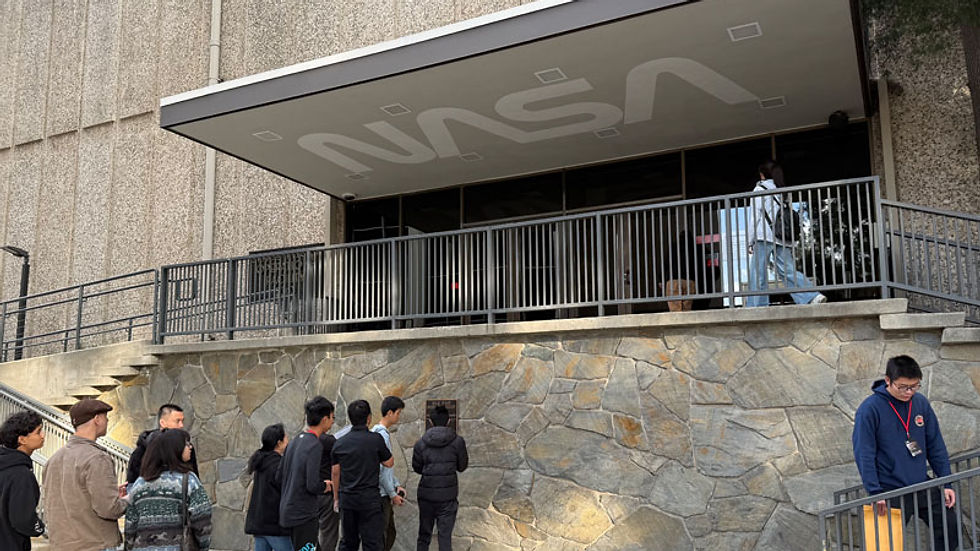 People queue outside a NASA building, standing by a stone wall and railing under a gray awning with a large NASA logo.