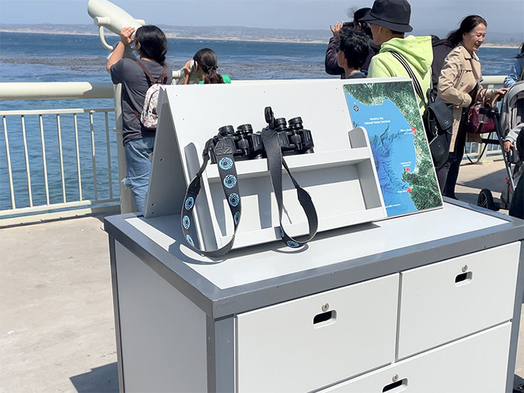 Binoculars and a map on a cabinet by a waterfront. People with cameras look out over the ocean, a bright and sunny day.