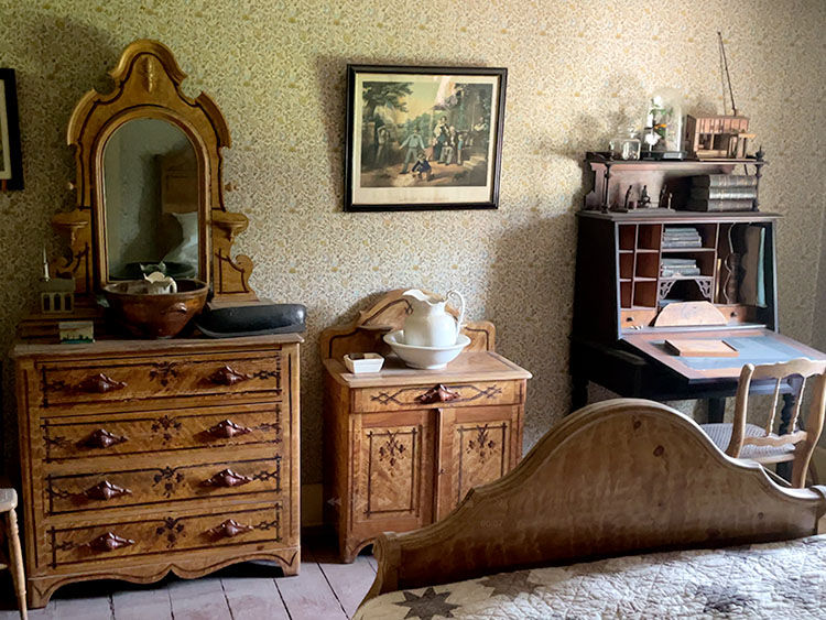 Vintage bedroom with ornate wooden dresser and mirror, floral wallpaper, framed painting, pitcher set, and a writing desk. Cozy, nostalgic feel.