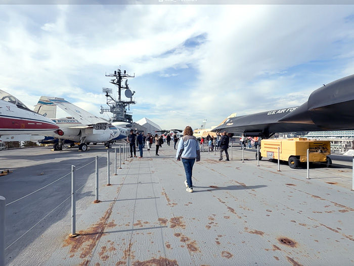 Visitors walk between fighter jets on an aircraft carrier deck under a cloudy sky.