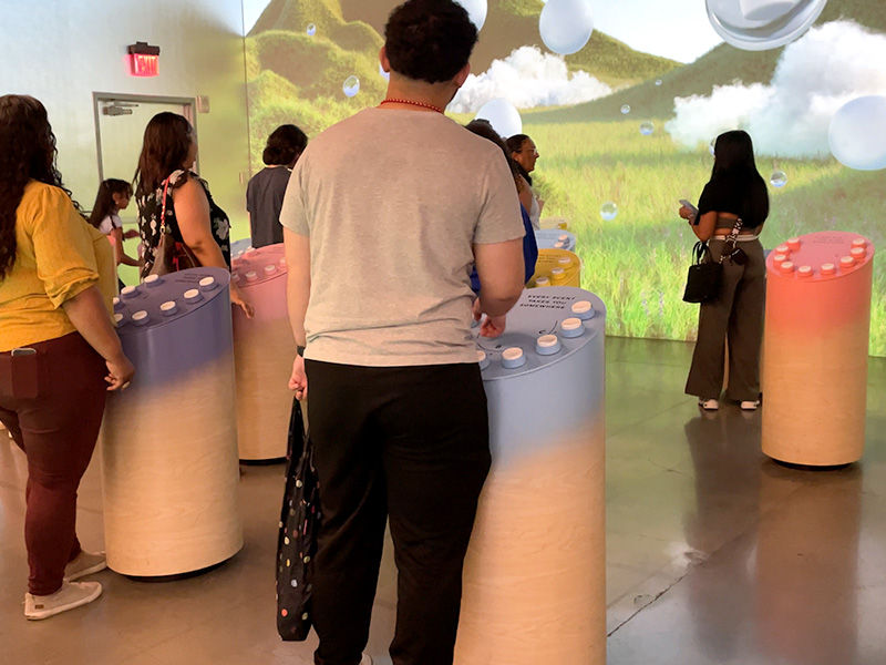 People interact with colorful cylindrical displays in a room with a nature mural backdrop. Mood is engaged; colors include blue and pink.