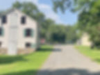 Historic village scene with a gravel path, old white wooden buildings, green shutters, and lush trees under a clear blue sky. Peaceful ambiance.