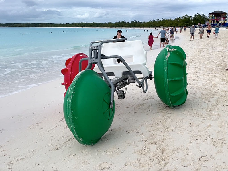 Large water tricycle with green wheels on a sandy beach, people walking in background by the blue ocean, cloudy sky above.