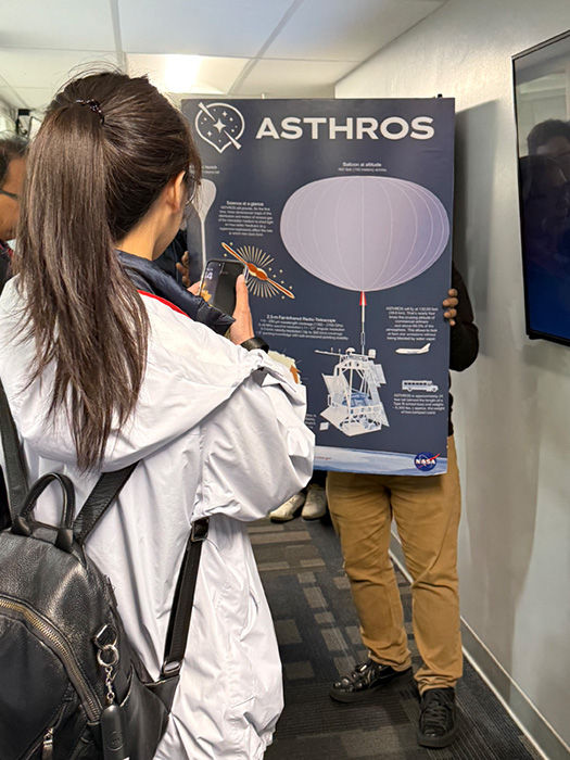 Person photographs a NASA ASTHROS poster in a hallway, showing mission graphics and text while another person holds it up.