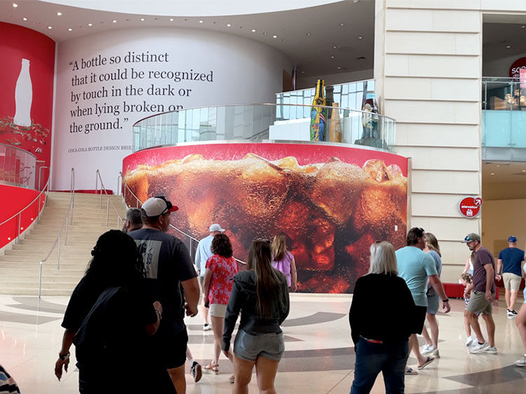 People walk through a Coca-Cola museum lobby with a giant soda mural. Red accents and a quote about bottle design are visible on the walls.