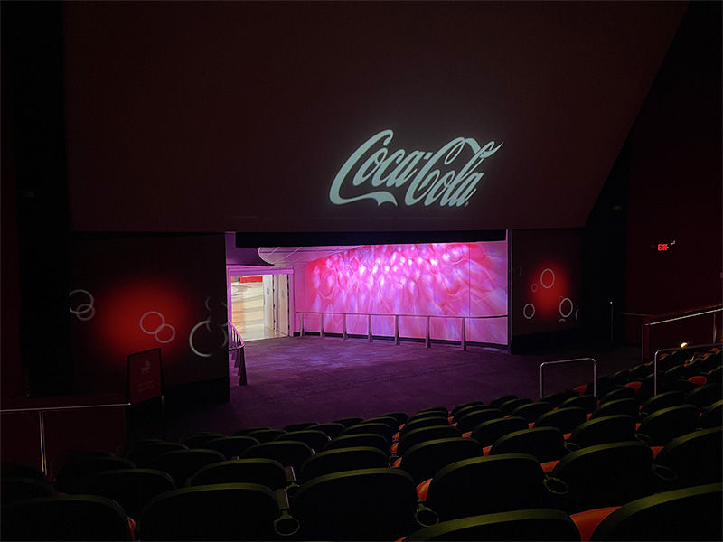 Dimly lit theater with empty seats, pink-lit walls, and a "Coca-Cola" projection on the screen, creating a calm and inviting mood.
