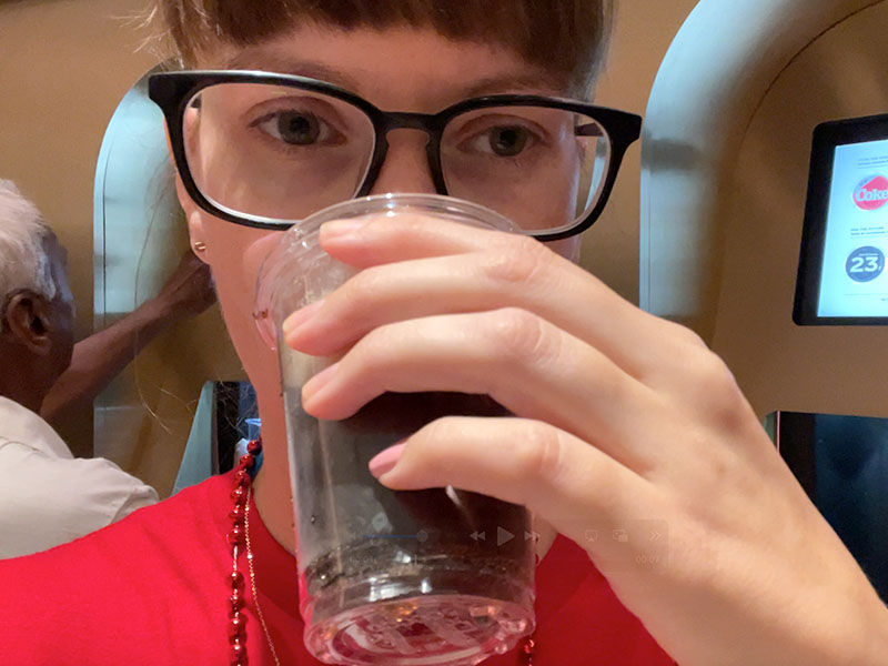 Person in glasses drinks from a plastic cup in a cafe. Background shows another person and a screen with text. Mood is casual.