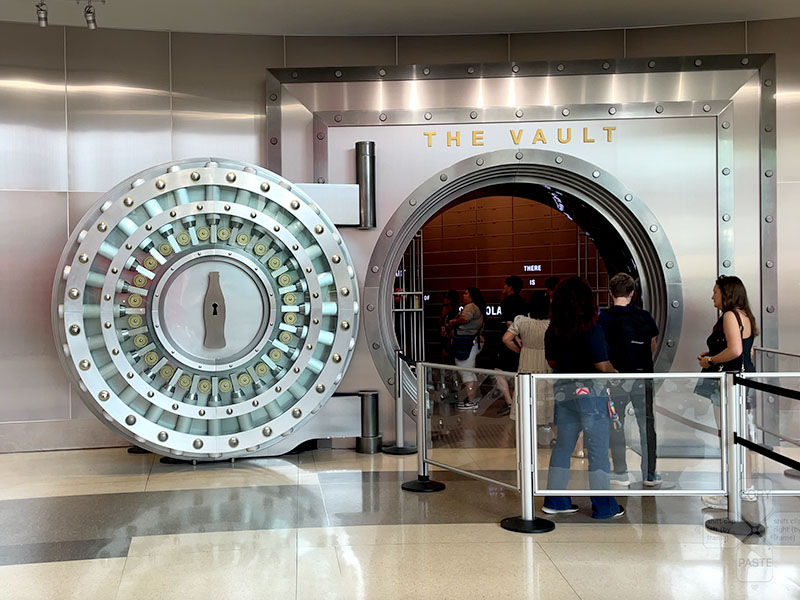 People enter a large metal vault labeled "The Vault." The door features a bottle design. The interior is dimly lit with a few visible signs.